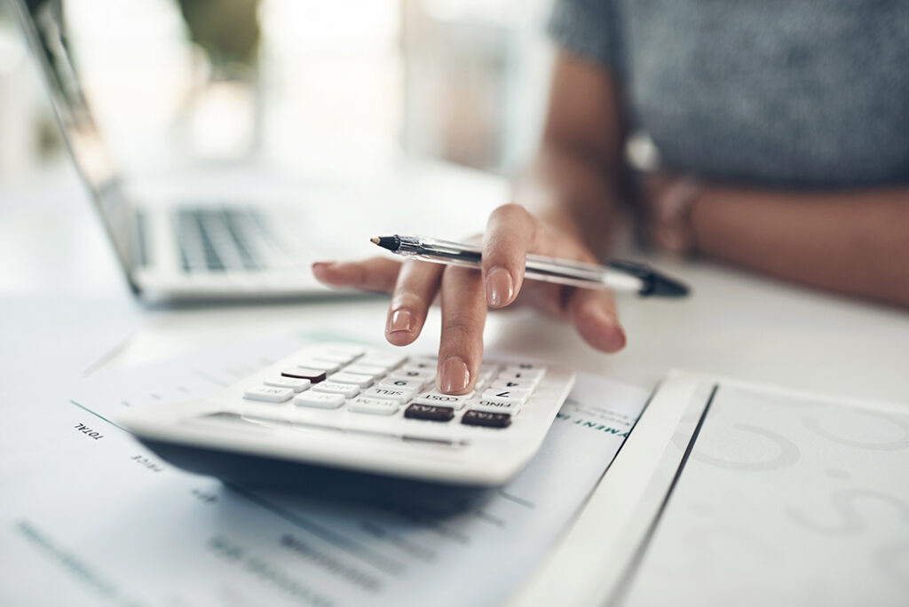 Close up view of woman's hand using calculator with pen and laptop.